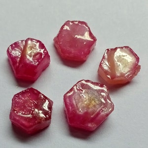 May include: Five faceted, pink ruby gemstones with a shiny, polished finish. The gemstones are arranged in a circle on a white surface.
