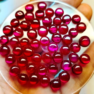 May include: A collection of round, red gemstones in a clear glass dish. The gemstones are various sizes and have a shiny, polished finish.