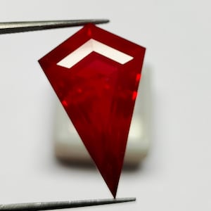 Puede incluir: Una gema roja vibrante, con forma de cometa y un recorte blanco en forma de cheurón. Sujeta con pinzas, mostrando su superficie pulida y diseño intrincado. La gema es probablemente para la fabricación de joyas.