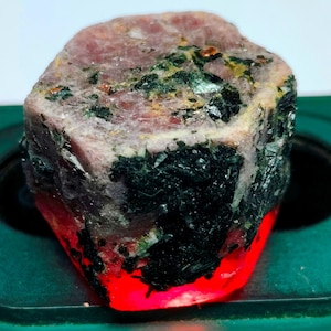 May include: A rough, uncut pink ruby gemstone with green inclusions. The ruby is illuminated from below, causing it to glow red.