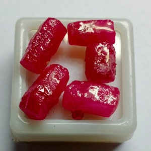 May include: Five rough, pink ruby gemstones in a white plastic container. The gemstones are various sizes and shapes.