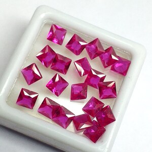 May include: A collection of square-cut, vibrant pink gemstones displayed in a white tray. The gemstones are faceted, reflecting light and showcasing their rich color. The arrangement highlights the precision of the cuts and the brilliance of the stones.