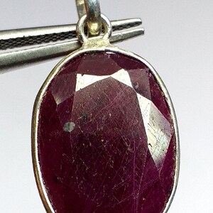 Może przedstawiać: Srebrny wisiorek z dużym, owalnym, fasetowanym rubinem. Rubin jest ciemnoczerwony i ma lekko fakturowaną powierzchnię.