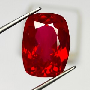 May include: A faceted, cushion-cut ruby gemstone. The gem is a deep, vibrant red with internal reflections. Held by silver tweezers against a white background, the ruby's facets catch the light, creating a brilliant display of color and sparkle. A close-up view of a precious stone.