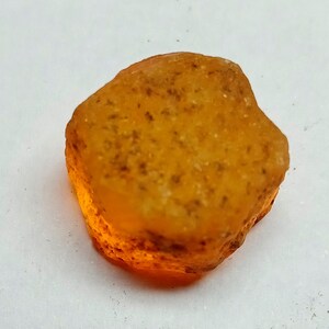 Op de afbeelding: Een ruwe, oranje-bruine edelsteen met een doorschijnende, licht troebele uitstraling. De steen heeft een onregelmatige vorm en een glad, gepolijst oppervlak.