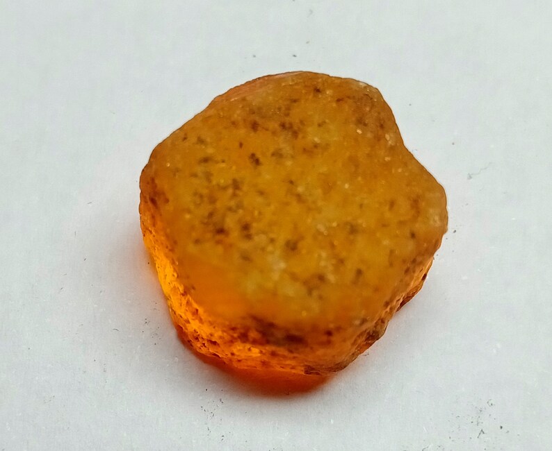 Op de afbeelding: Een ruwe, oranje-bruine edelsteen met een doorschijnende, licht troebele uitstraling. De steen heeft een onregelmatige vorm en een glad, gepolijst oppervlak.