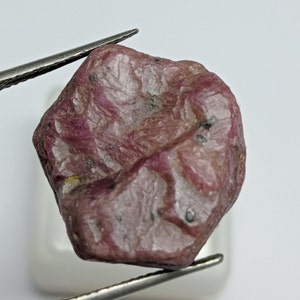 Può includere: Una pietra preziosa di rubino rosa grezza con una finitura naturale non lucidata. Il rubino ha un colore rosa intenso con inclusioni bianche e grigie.
