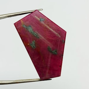 May include: A pink gemstone with a faceted, irregular shape. The gemstone has a dark red colour with white and grey streaks throughout.