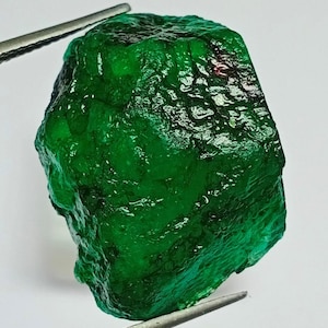 May include: A large, rough, green emerald gemstone with a slightly reddish hue. The emerald is faceted and has a glossy finish.
