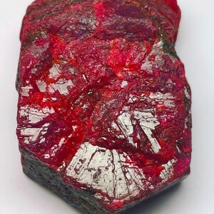 Puede incluir: Una gema de rubí en bruto y sin cortar, de color rojo intenso. La superficie tiene un aspecto texturizado con áreas de estructuras cristalinas blanco plateado. El rubí tiene forma irregular, mostrando su forma natural y en bruto. La gema está sobre un fondo blanco.