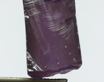 May include: A rectangular, translucent, dark purple gemstone held by metal tweezers. The gemstone has a rough, textured surface with visible striations. The background is plain white, highlighting the gem's color and form.