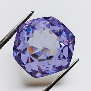 May include: A cushion-cut lavender gemstone with a faceted surface held by metal tweezers. The stone displays a translucent purple hue with internal light reflections and sharp geometric edges, showcasing a clear, polished, and symmetrical appearance.