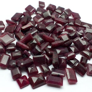 Könnte beinhalten: Ein Haufen loser, facettierter, dunkelroter Rubin-Edelsteine. Die Steine sind rechteckig und haben eine polierte Oberfläche.