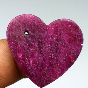 May include: A heart-shaped ruby gemstone with a deep pink hue. The gemstone has a rough, textured surface and a small hole drilled through the top.