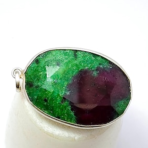 Puede incluir: Un colgante de piedra preciosa facetada con un bisel plateado. La piedra preciosa presenta una llamativa combinación de tonos rojo intenso y verde vibrante. El colgante es de forma ovalada y tiene un lazo para una cadena.