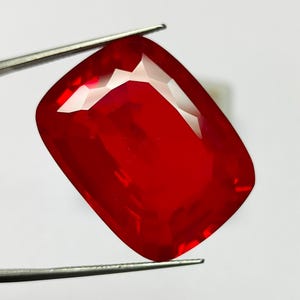 May include: A large, faceted, rectangular-shaped red gemstone is held by a pair of silver tweezers. The gem has a brilliant, reflective surface, showcasing multiple facets that catch the light. The gemstone is a vibrant, deep red color.
