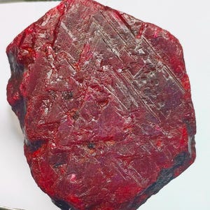 May include: A large, rough, red ruby gemstone. The ruby has a deep red color and a rough, unpolished surface. The ruby is a natural gemstone and has a unique, irregular shape.
