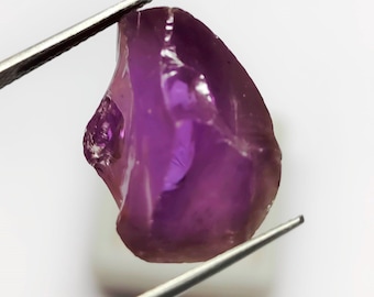 May include: A raw, uncut amethyst gemstone, held by tweezers against a white background. The gem is a deep purple color with a translucent quality, showcasing its natural, irregular shape. The tweezers are silver.