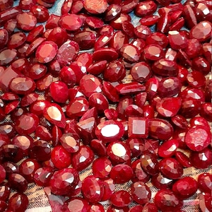 May include: A large collection of red gemstones, likely rubies, of various sizes and shapes. The gemstones are displayed on a patterned fabric background.