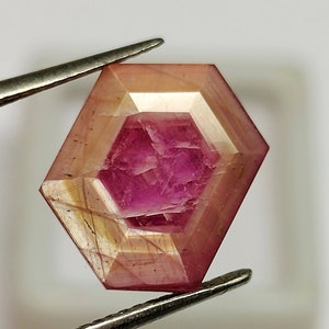 May include: A pink and white gemstone with a hexagonal cut. The gemstone is held by tweezers.