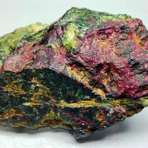 Puede incluir: Una roca rugosa de forma irregular con una mezcla de tonos rojo oscuro, verde y dorado. La roca tiene un aspecto estratificado con una variedad de texturas.