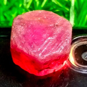May include: A large, faceted, pink ruby gemstone with a hexagonal shape. The ruby is sitting on a black surface.