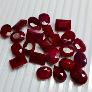 May include: A collection of red ruby gemstones of various shapes and sizes. The gemstones are arranged on a white surface.