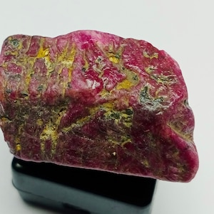 May include: A rough, unpolished ruby gemstone with a deep red color and a mix of dark red and gold inclusions. The ruby is sitting on a black surface.