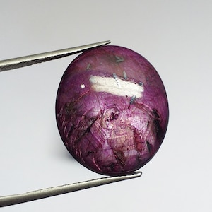 May include: A close-up of a purple oval-shaped gemstone with silver flecks and a shiny finish. The gemstone is held by tweezers.
