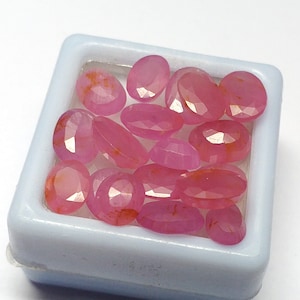 May include: A collection of pink oval-shaped gemstones in a white square container. The gemstones have a faceted cut and are translucent.