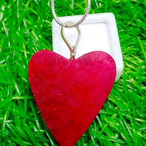 May include: A heart-shaped pendant in a deep red hue, suspended from a silver chain. The pendant has a textured surface and is set against a backdrop of vibrant green grass.