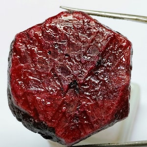 May include: A deep red ruby gemstone with a hexagonal shape and a smooth, polished surface. The ruby has a dark red colour with a slight purple hue. The gemstone is unfaceted and has a natural, rough edge.