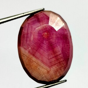 Pode incluir: Gema de rubi oval e facetada. A gema exibe um tom vermelho-rosado profundo com áreas mais claras e translúcidas. As facetas criam um padrão geométrico, e a pedra é segurada por uma pinça prateada contra um fundo branco.