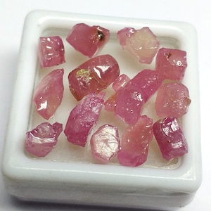 Op de afbeelding: Een verzameling ruwe roze robijn edelstenen in een witte vierkante container. De ruwe edelstenen zijn in verschillende vormen en maten.