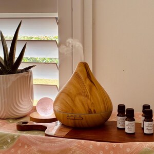 May include: A teardrop-shaped essential oil diffuser with a light wood grain finish emitting a thin stream of mist. Beside it are three small amber glass bottles with black caps and white labels, a round pink crystal, and a green succulent in a white pot.