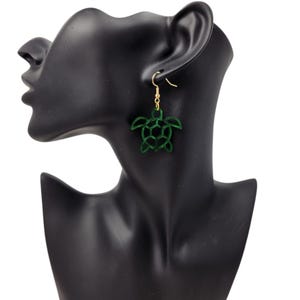 May include: Dark green turtle-shaped earrings with gold-coloured hooks. The earrings have a cutout design. The earrings are displayed on a black mannequin head.