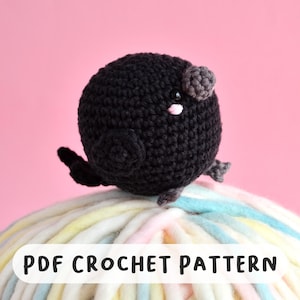 Może przedstawiać: Ręcznie robiony czarny szydełkowy ptak z szarymi akcentami, w tym małymi skrzydłami i uszami, siedzi na kłębie kolorowej przędzy. Ptak ma różowy nos i jest umieszczony na różowym tle. Tekst na przędzy brzmi "PDF CROCHET PATTERN."