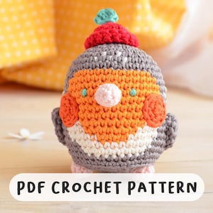 Może przedstawiać: Szydełkowany ptak z czerwonym kapeluszem, pomarańczową piersią i szarym korpusem. Ptak ma biało-szary wzorzysty brzuch, różowe stopy i różowy dziób. Tekst "PDF CROCHET PATTERN" jest wyświetlany na białym banerze u dołu.