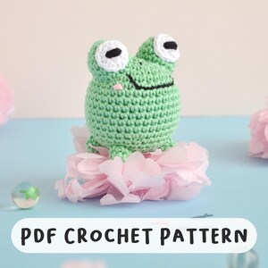 May include: A crocheted green frog with white and black eyes sits atop pink paper flowers. The frog has a smiling mouth and a small pink nose. The text "PDF CROCHET PATTERN" is displayed below the frog.