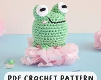 May include: A crocheted green frog with white and black eyes sits atop pink paper flowers. The frog has a smiling mouth and a small pink nose. The text "PDF CROCHET PATTERN" is displayed below the frog.