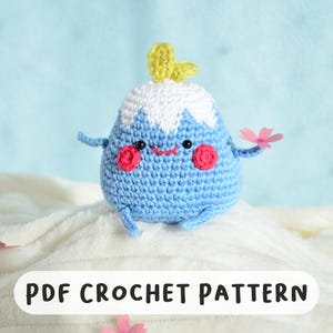 May include: A crocheted blue mountain character with a white snowy peak, rosy cheeks, and a smiling face. It has small arms and legs, and holds a pink flower. The text "PDF CROCHET PATTERN" is at the bottom.