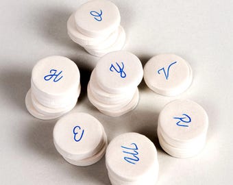 May include: White, round, stacked paper coasters with blue handwritten letters. The coasters are arranged in a circular pattern on a light gray surface. Each stack has a different letter.