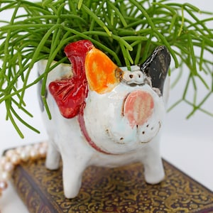 May include: A white ceramic planter shaped like a dog with red, orange, and black accents. The planter is holding a green succulent plant.