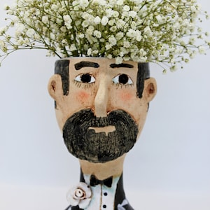 Pode incluir: Um vaso de cerâmica em forma de cabeça de pessoa com bigode e smoking. O vaso tem uma flor branca na lapela e está cheio de flores brancas de gipsofila.