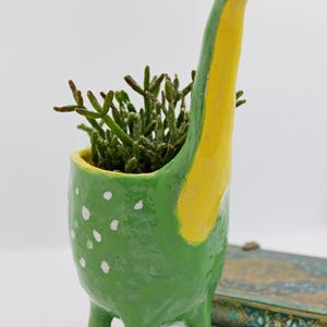 Peut inclure: Un pot de fleurs en céramique peint à la main en forme de dinosaure, aux couleurs vert et jaune, avec des pois blancs. Le pot en forme de dinosaure contient une petite plante succulente verte. Le pot a trois pattes et un long cou.