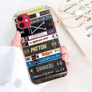 Puede incluir: Una funda transparente para teléfono con un collage de cintas VHS vintage. Las cintas muestran títulos de películas como "Patton" y "Schindler's List". La funda tiene un módulo de cámara rojo y la sostiene una persona.