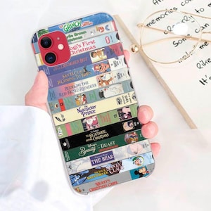 May include: A clear phone case printed with a collage of VHS tape spines. Titles such as "The Grinch," "Beauty and the Beast," and "Mickey's Christmas Carol" are displayed. The case is held in a person's hand.