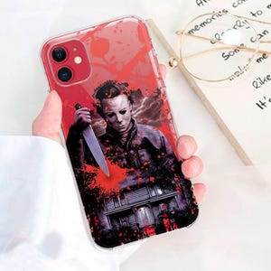 May include: A clear phone case with a red, white, and black graphic of a man holding a knife. The graphic is a horror movie character with blood splatters on the case.