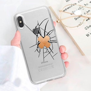 Puede incluir: Una funda transparente para teléfono con un diseño de pantalla agrietada. Un pequeño vendaje marrón cubre la grieta. La funda tiene la palabra "iPhone" impresa en la parte posterior.