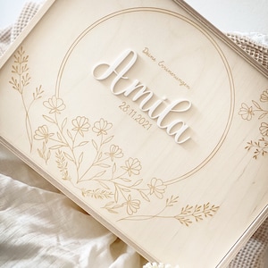 Puede incluir: Una caja de madera con fondo blanco y dise&ntilde;os florales dorados. La caja tiene un c&iacute;rculo en el centro con el nombre "Amilia" y la fecha "28.11.2021" escritos en blanco. El texto "Deine Erinnerungen" est&aacute; escrito en dorado sobre el nombre.